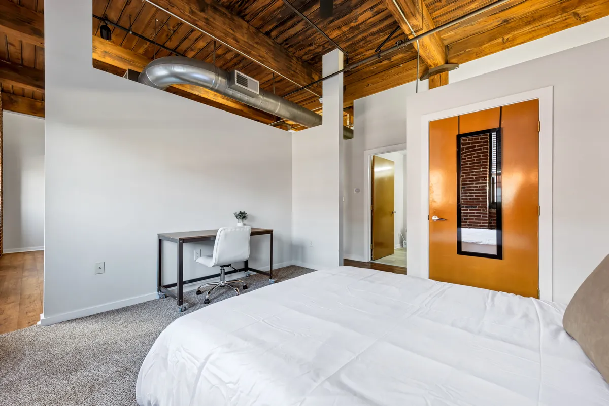 A spacious bedroom with a comfortable bed, a dedicated desk and chair for work or study, and warm exposed beams overhead. The scene includes an orange door leading toward the adjoining bathroom, combining relaxation with functional workspace—all within a calm, contemporary setting.