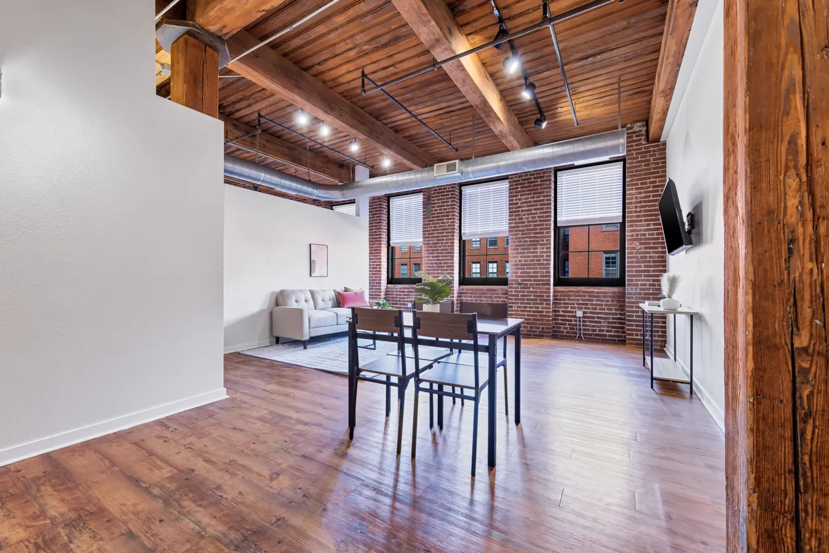An open-plan space featuring visible wooden beams, an exposed brick wall, a cozy sofa, and a well-appointed dining table with chairs. This setting balances areas for relaxation and socializing, with both the living and dining areas flowing seamlessly into one another under high ceilings—emphasizing the open-concept living described in the section.