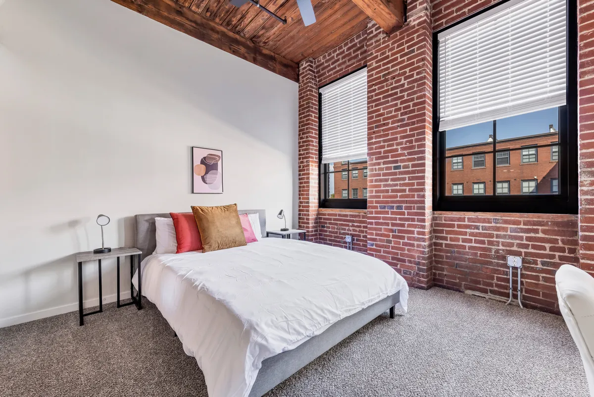 A serene bedroom featuring a plush bed with high-quality linens, bathed in natural light from expansive windows. The space is enhanced by a calming color palette, a stylish brick accent wall, elegant artwork, and a cozy nightstand. The overall ambiance is tranquil, with soft textures and thoughtful details providing a peaceful retreat.