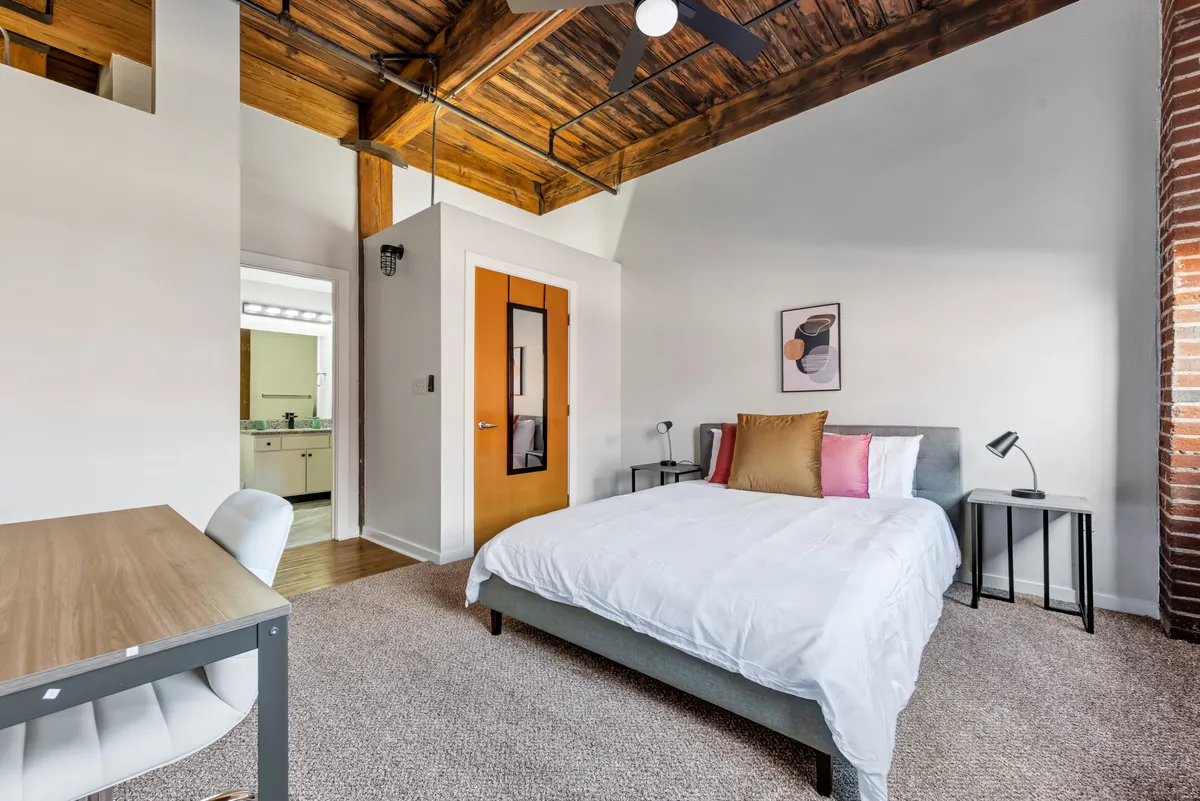 The image showcases a stylish bedroom with a comfortable bed adorned with decorative pillows, exposed wooden ceiling beams that add historic charm, a modern desk and chair ideal for remote work, and tasteful artwork on the wall. Natural light fills the space, creating a warm and inviting atmosphere.