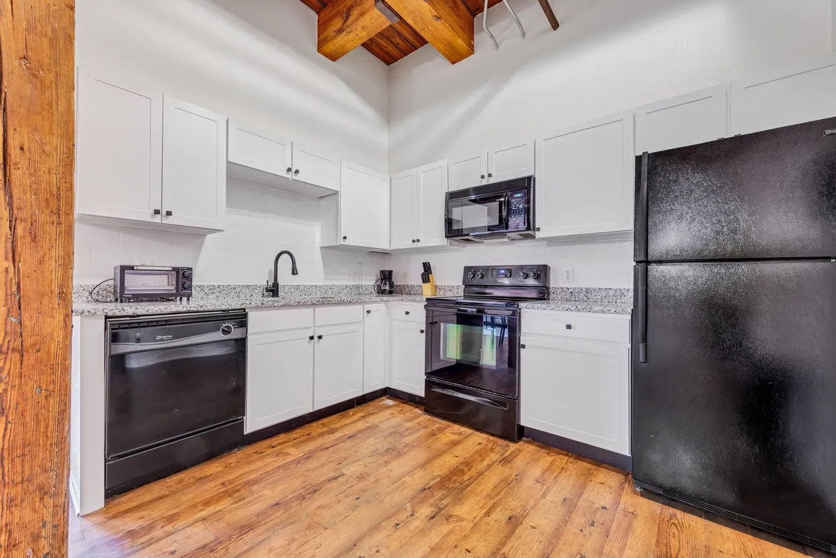 A modern kitchen view with granite countertops and sleek black appliances, accented by wooden ceiling beams. The space is set up for active meal preparation and entertaining guests, emphasizing the stylish, contemporary design and practicality for hosting gatherings or preparing family meals.