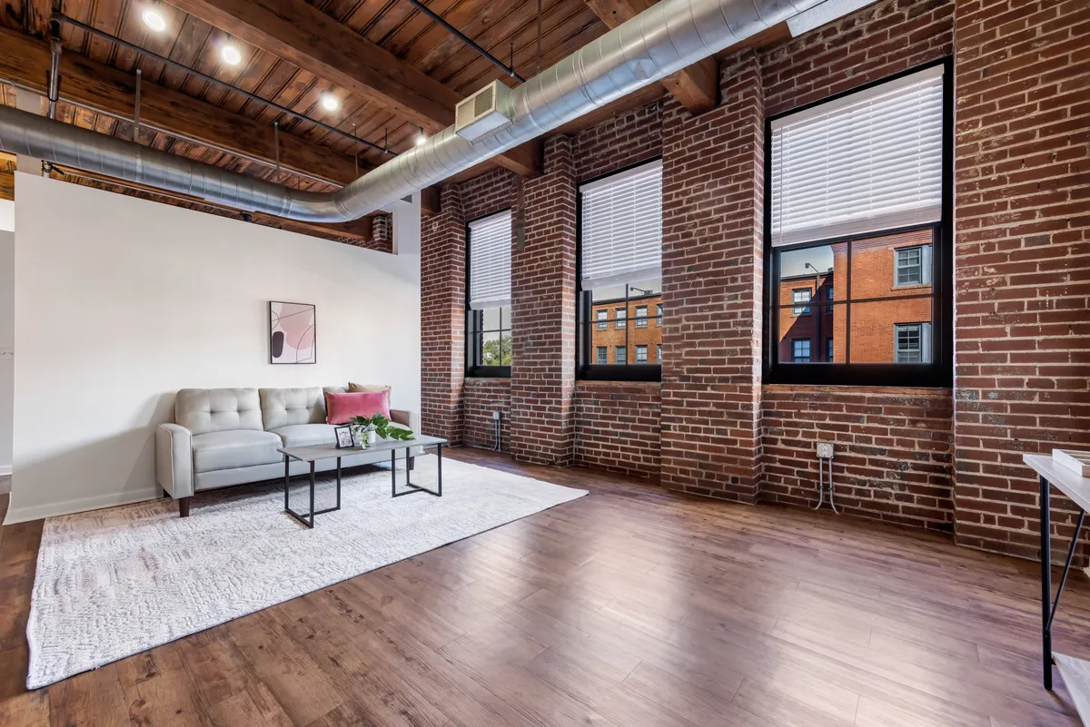 A spacious living area with a plush sofa and coffee table, set against a striking exposed brick wall. Large windows allow abundant natural light to pour into the room, illuminating the contemporary décor and historic architectural elements. The warmth of the brick and wood materials creates an inviting and comfortable ambiance, blending modern furnishings with preserved character.