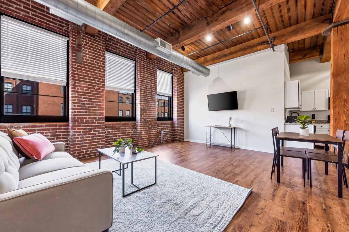 A bright, contemporary loft space with a comfortable sofa, sleek coffee table, expansive windows letting in natural light, a dining area, and a TV. The layout offers several zones for sitting, relaxing, or focusing on tasks, all in a setting that feels both modern and homelike.
