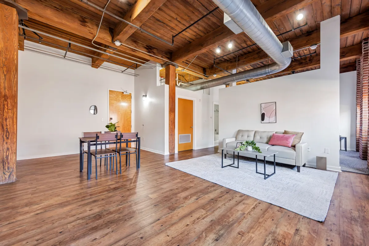 A welcoming loft interior featuring exposed wooden beams overhead, a plush sofa tucked into a cozy corner, and a large dining table nearby. The atmosphere is intimate and inviting, with soft lighting that suggests both relaxation and productivity. The space is designed to accommodate various activities, from socializing to reading or working.