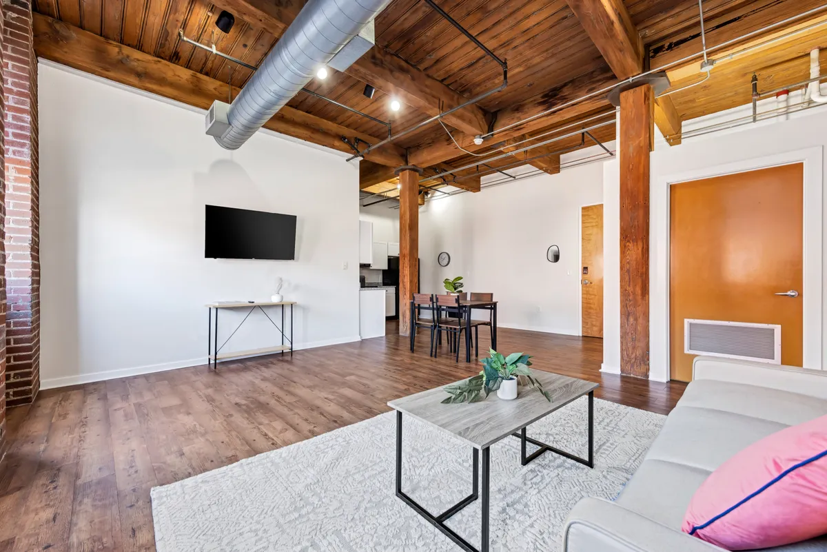 A spacious, modern living area featuring a comfortable sofa, stylish coffee table, TV, and dining table beneath exposed wooden beams. The setting feels open and inviting, with plenty of room for relaxing or entertaining after a day out in the city.